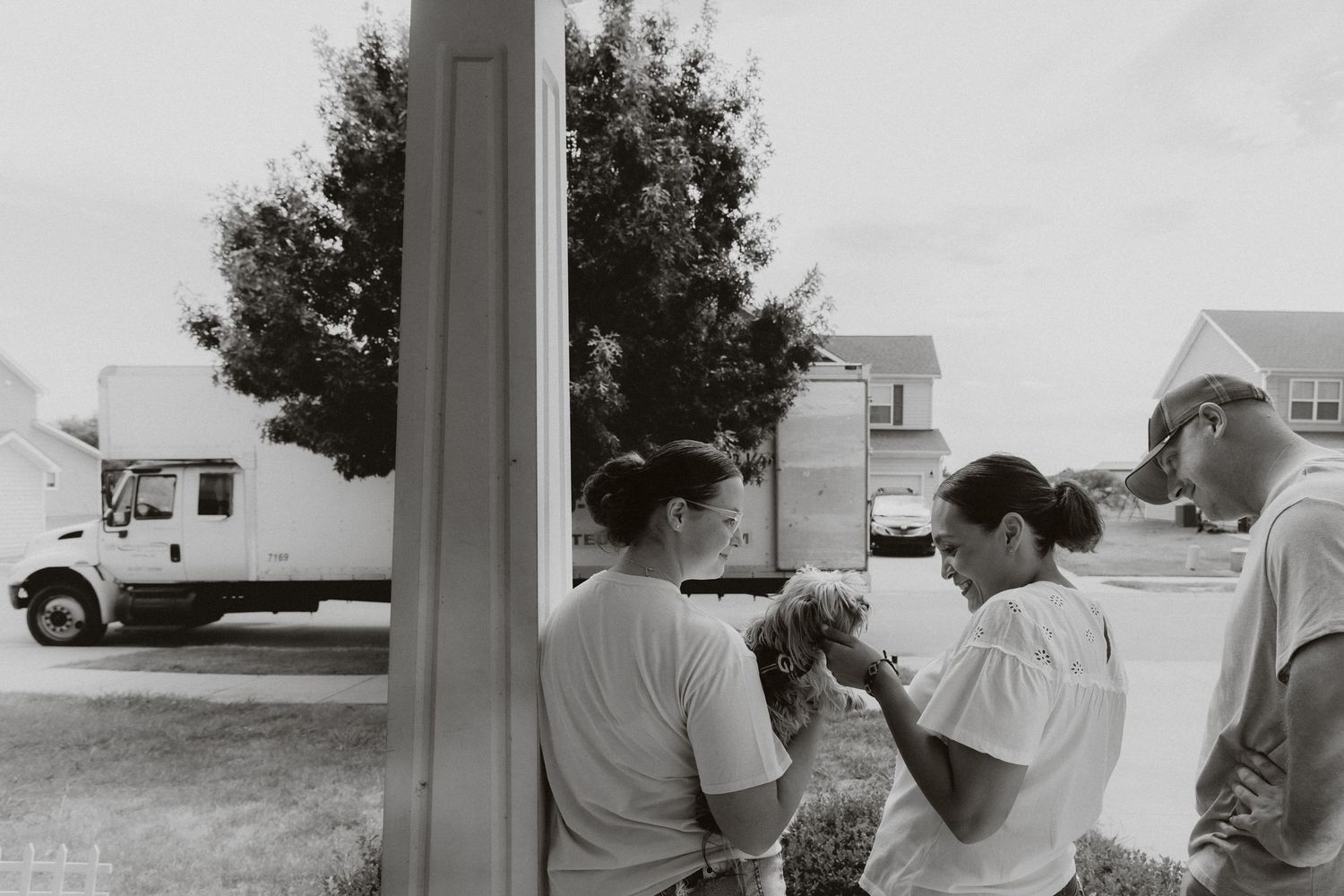 Black and white photo of three people examining something outside a home with a moving truck in the background.