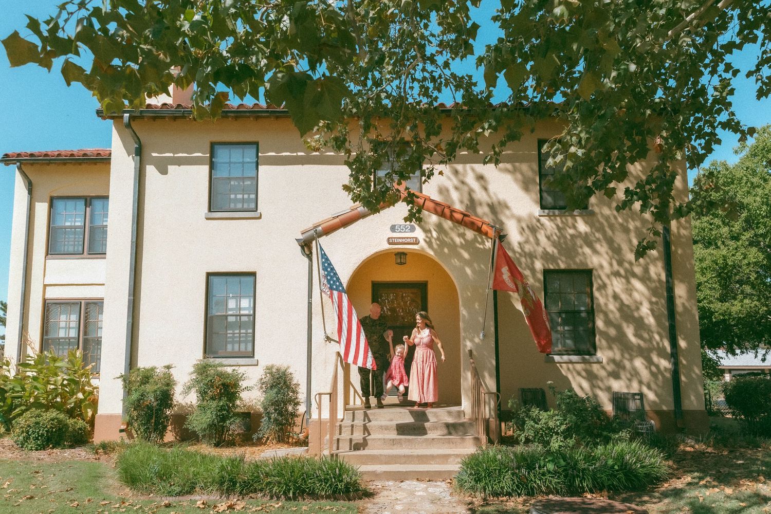 Two-story Mediterranean-style building with American flag, surrounded by trees, as a woman in pink dress exits.