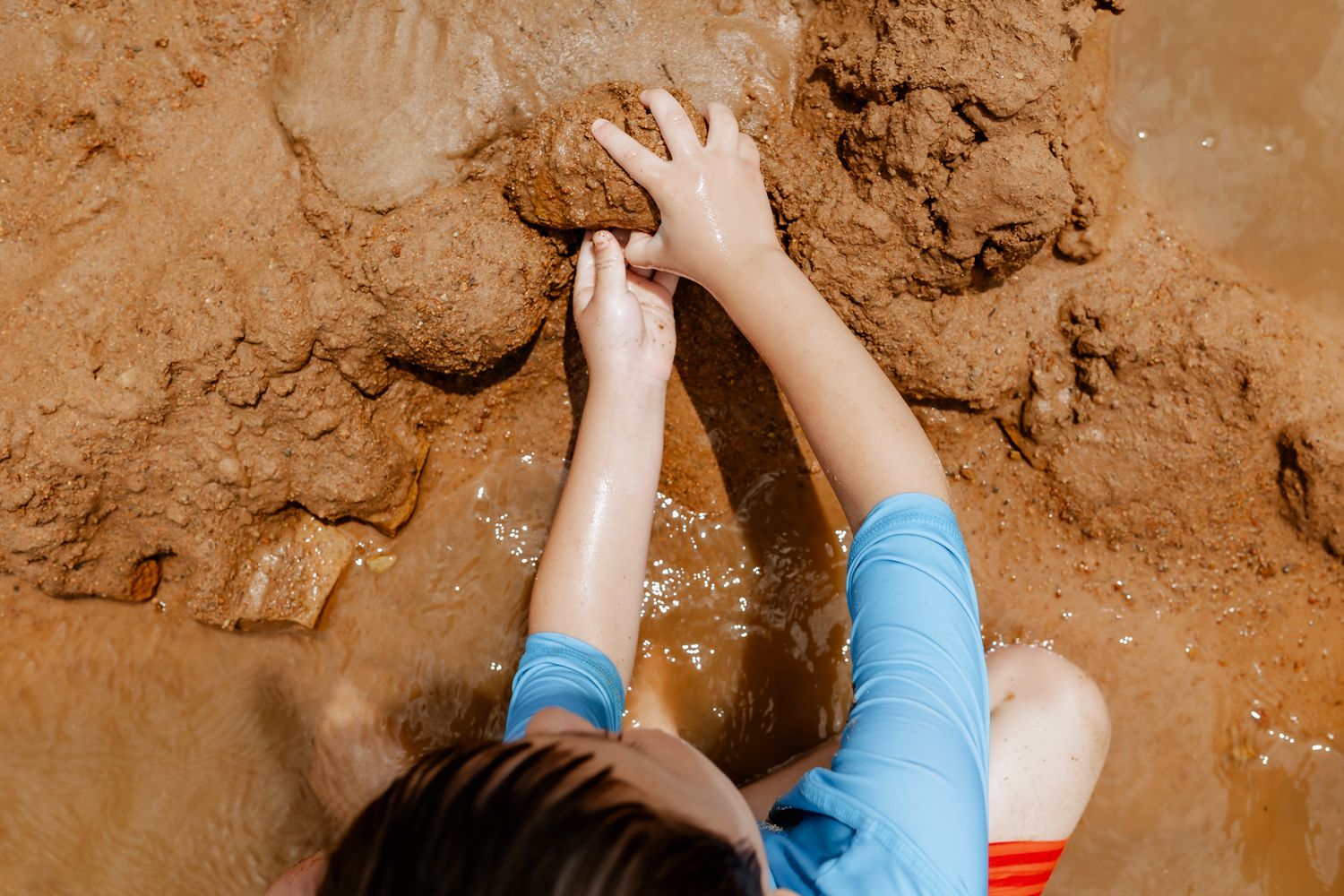 Hands reach down to dig and play in wet brown sand at the beach.
