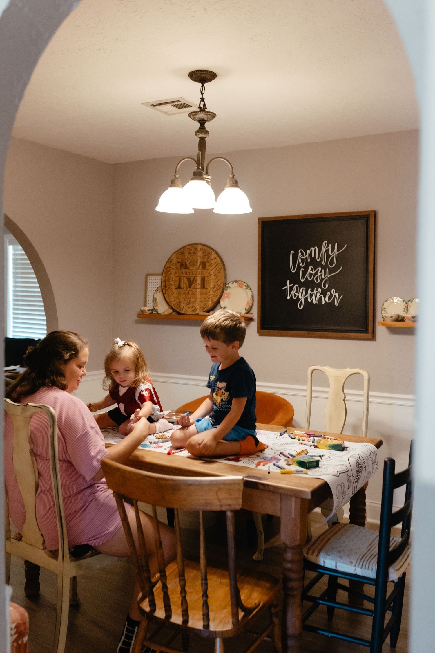 A mother and two young children sit together at a wooden dining table crafting in a cozy home dining room.