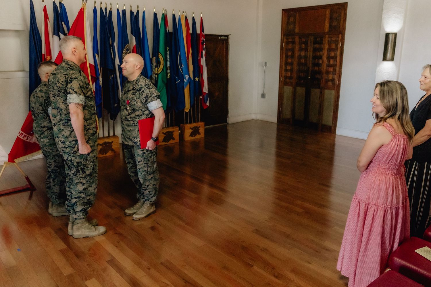 Marines in uniform stand before colorful flags during a military ceremony indoors, with civilians observing nearby.
