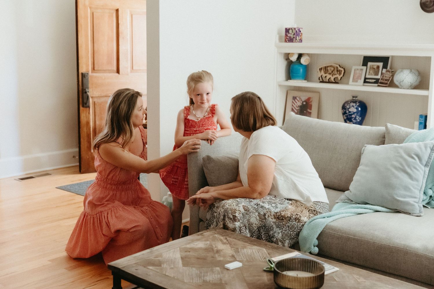 Two women interact with a young girl in a bright living room with hardwood floors and a gray sofa.