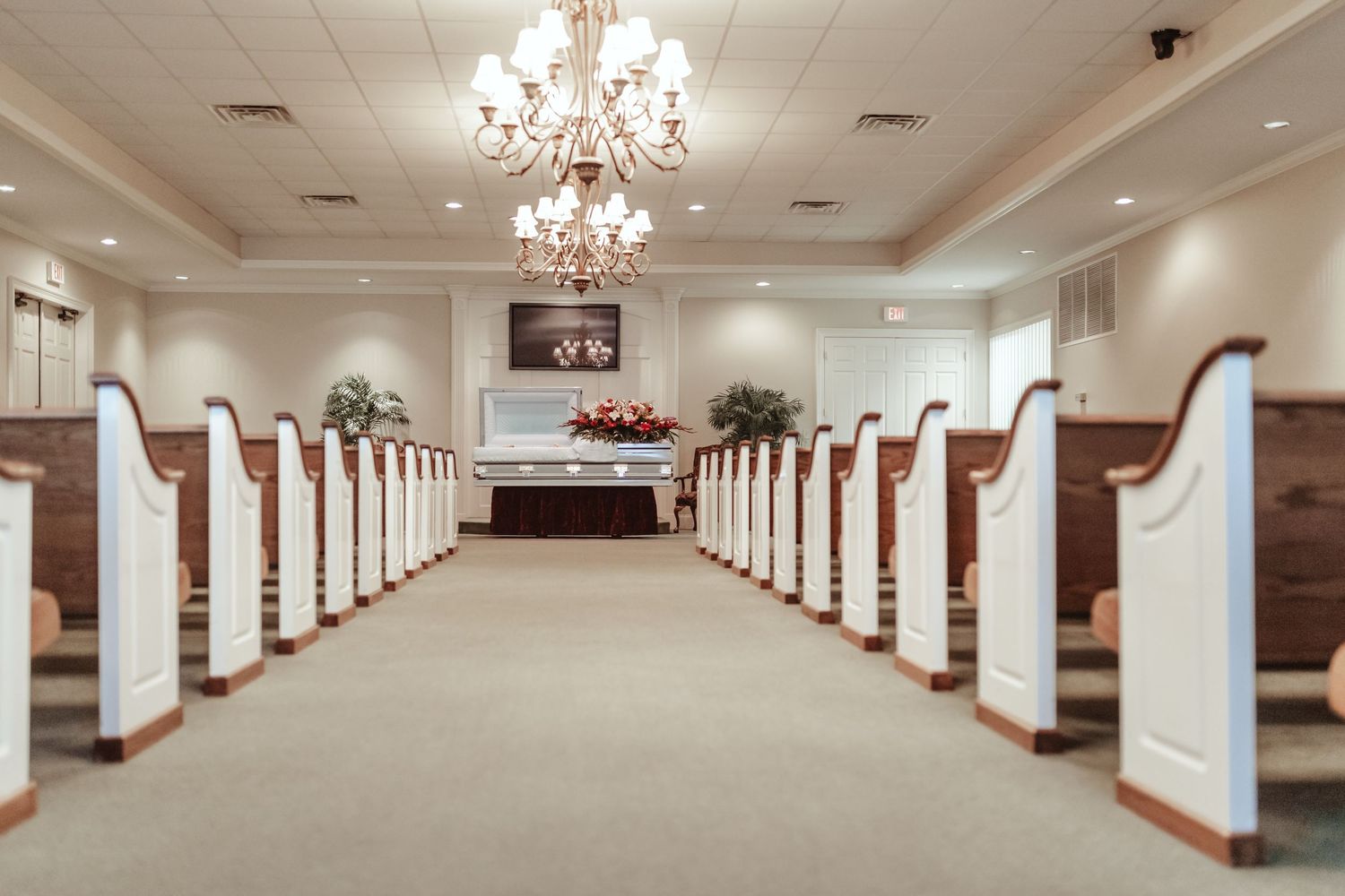 Long rows of white church pews lead to an altar under crystal chandeliers in a funeral home chapel.