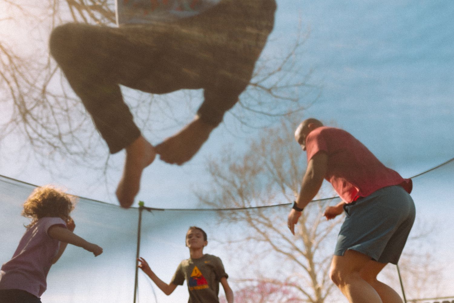 Children jumping on a backyard trampoline on a sunny day, viewed from below against a blue sky.