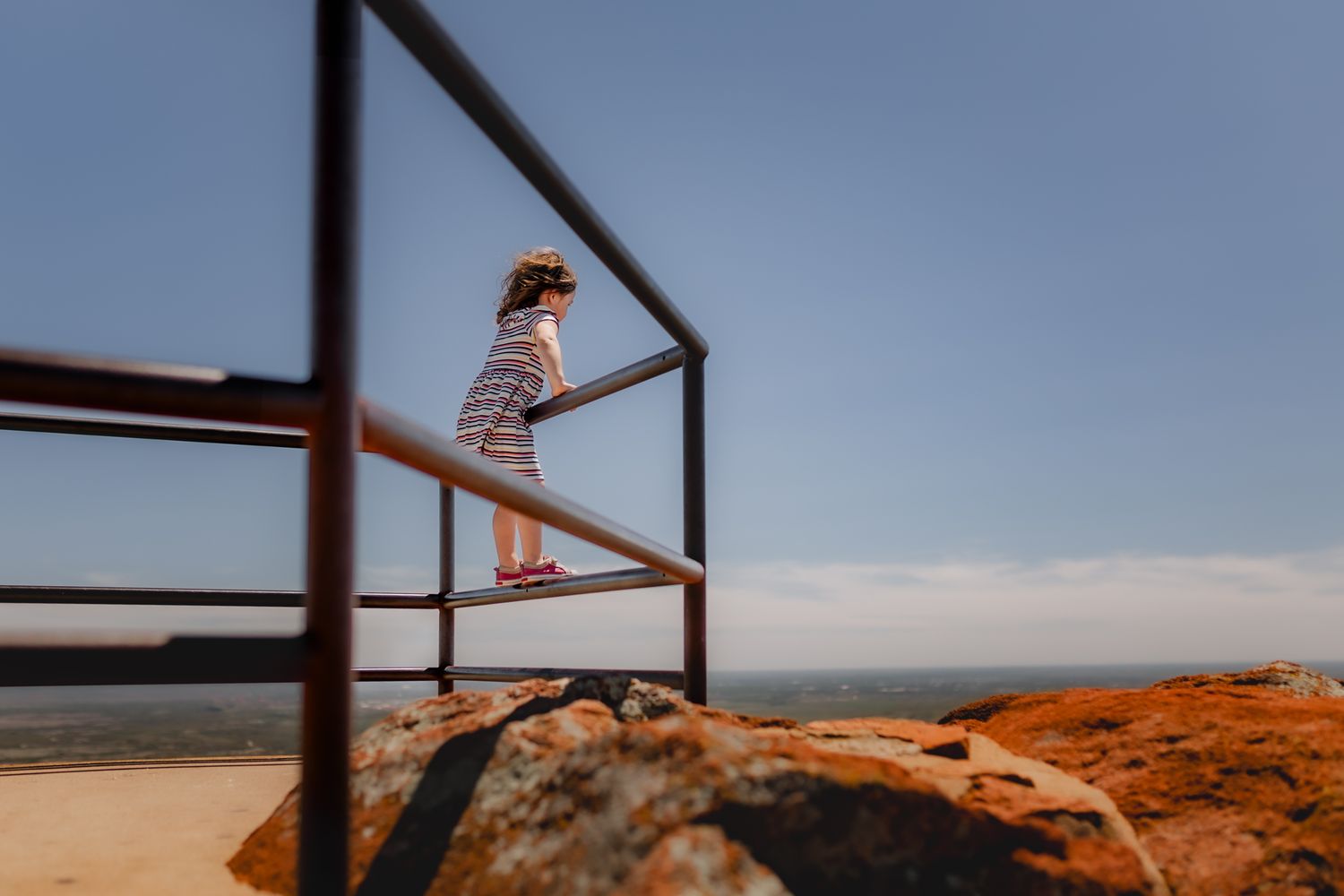 Figure standing on rusty railing overlooking scenic coastal vista on sunny day.