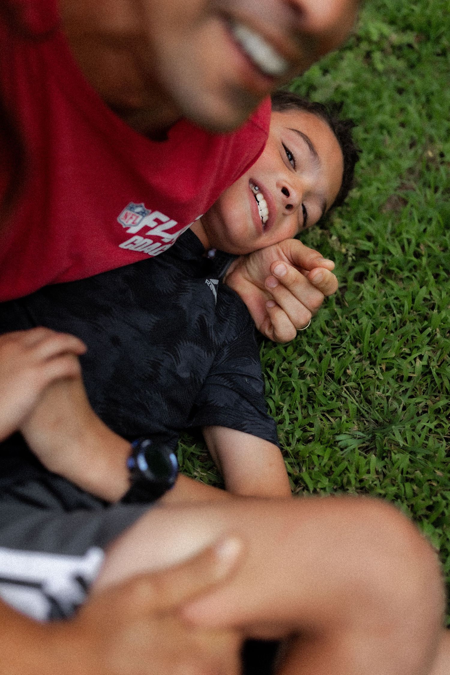 A smiling child lies on green grass while an adult in a red NFL shirt leans over them playfully outdoors.