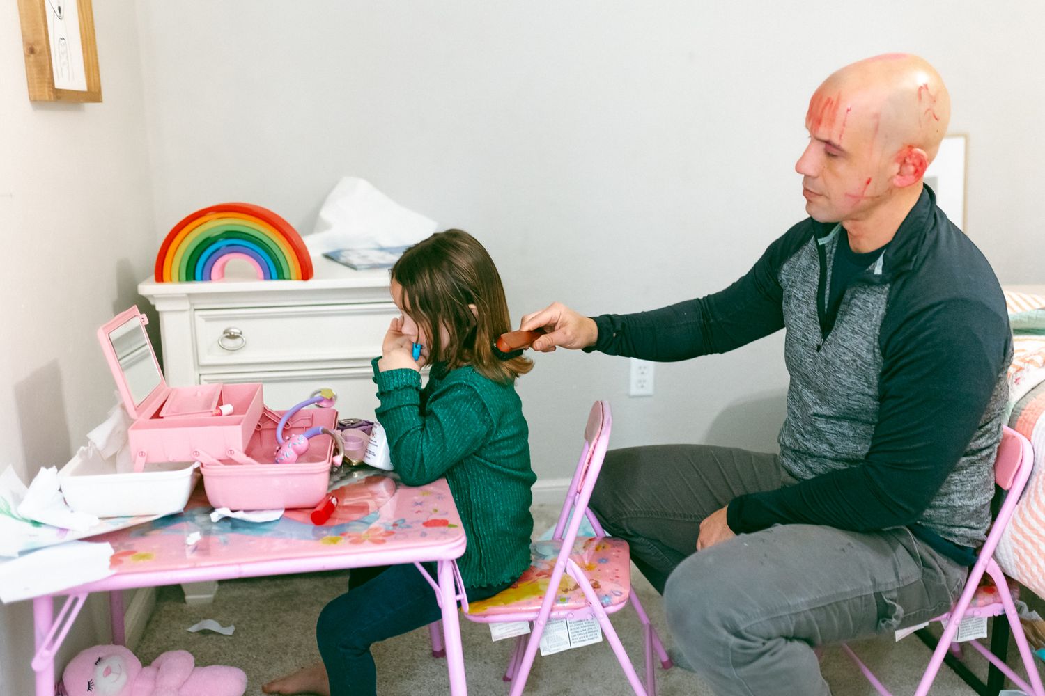 A parent helps their child with homework at a pink folding table with a rainbow decoration on the wall.