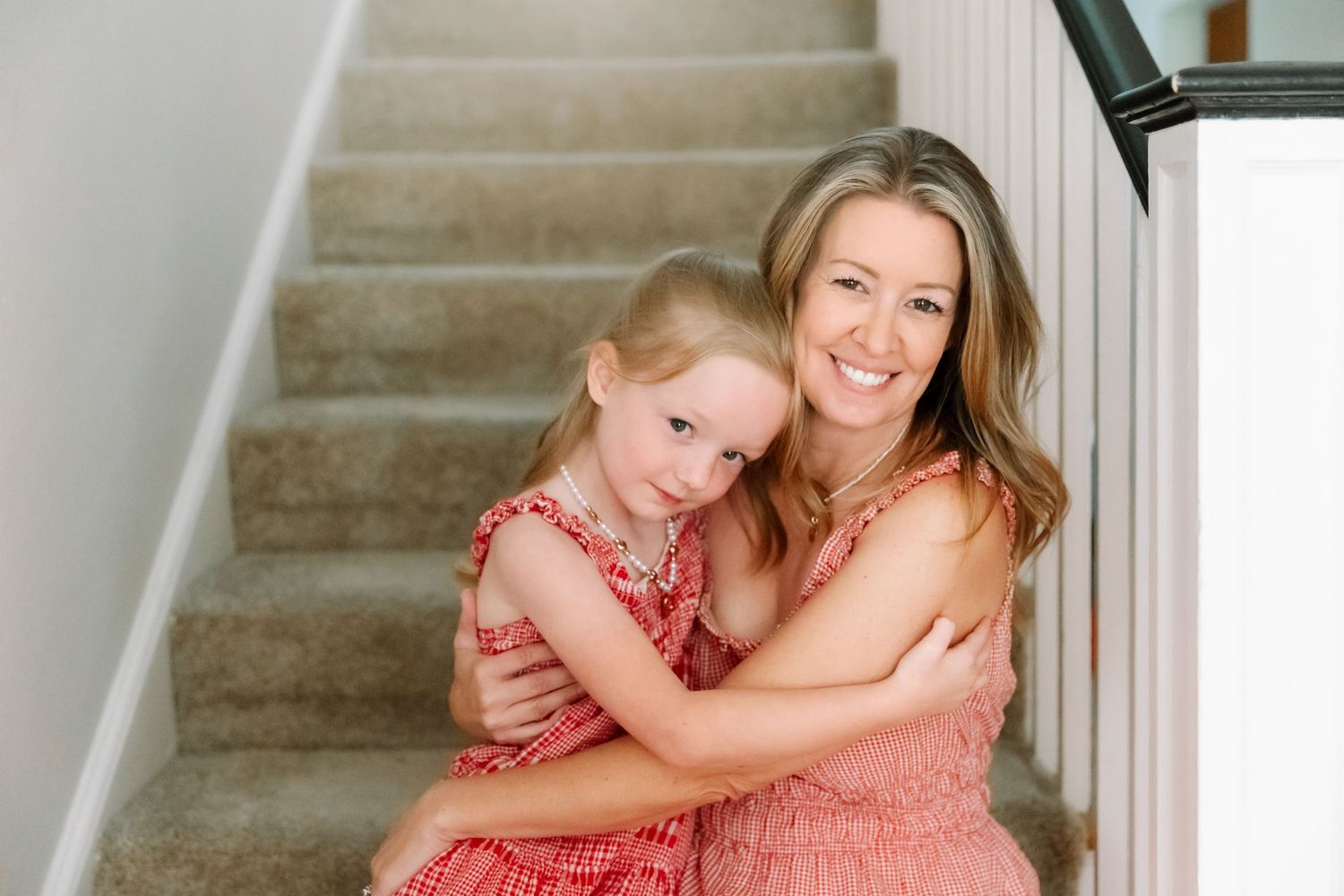 A smiling mother and young daughter in matching pink dresses sitting on carpeted stairs hugging.