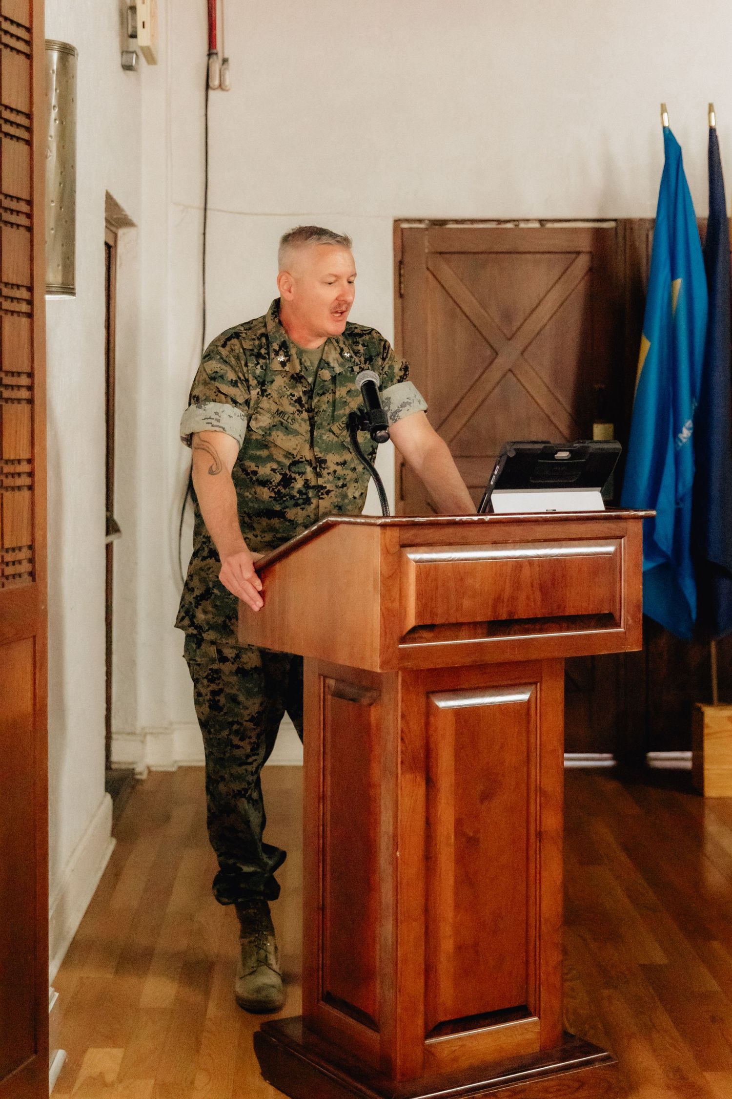 Military officer in camouflage uniform speaking at a wooden podium with flags in the background.