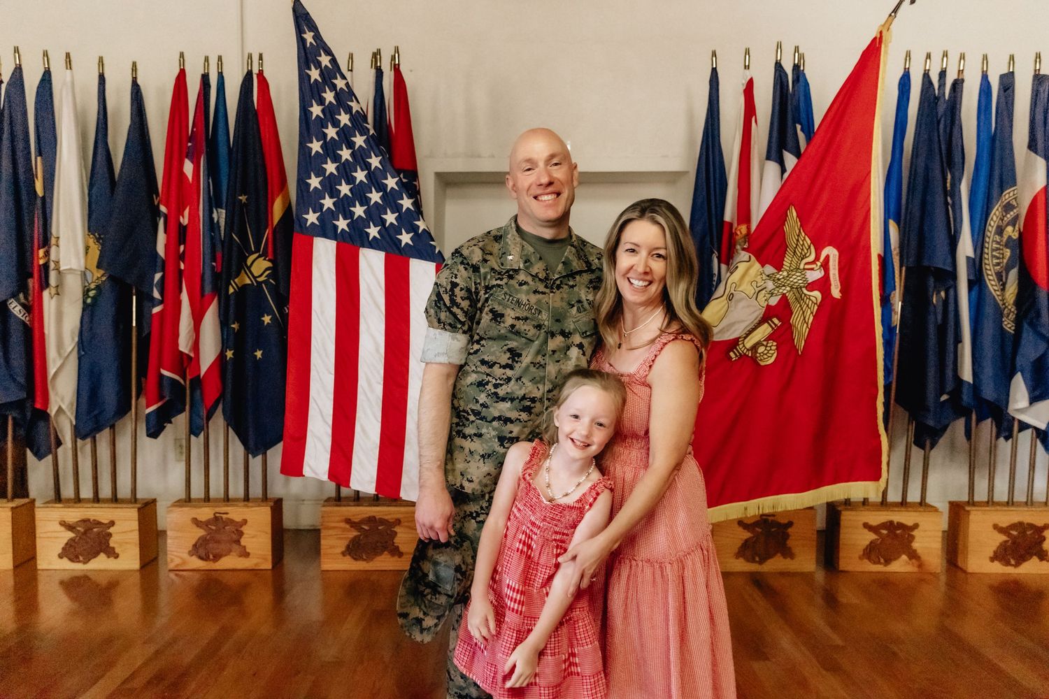 Military family portrait with soldier in camouflage, wife in pink dress, and young daughter before American flags.