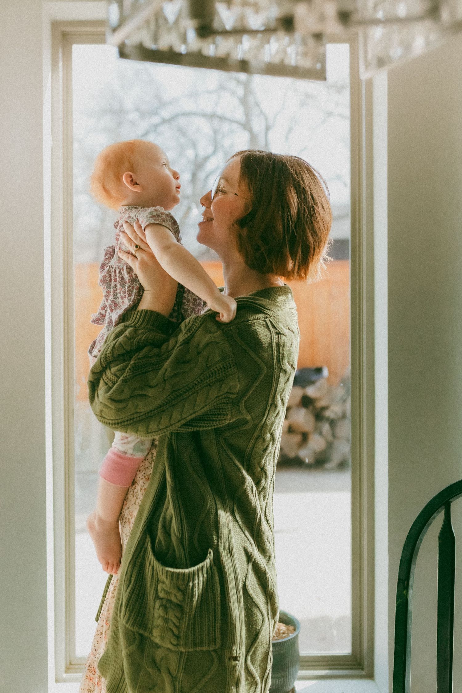 A parent holds a baby up in the air near large windows in a sunlit room while wearing an olive green jacket.