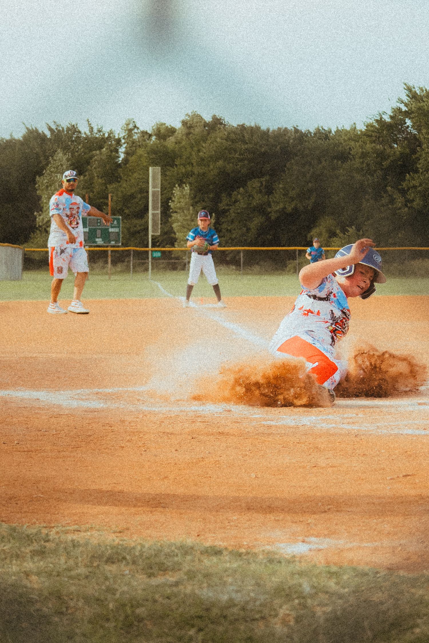 Baseball player slides into home plate in a cloud of dirt during a game, with teammates visible in the background.