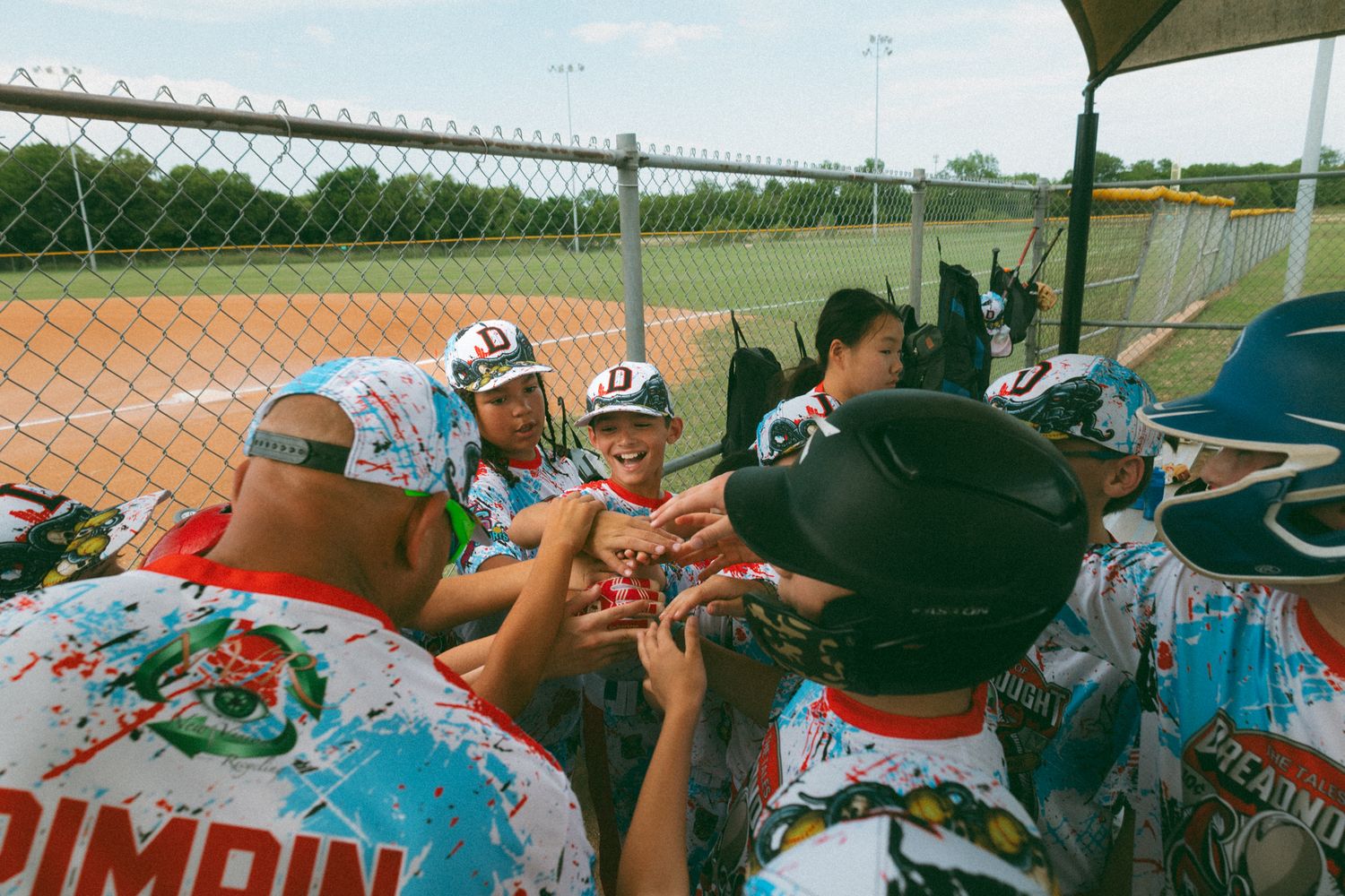 Youth baseball team in colorful Primpin uniforms huddling together in dugout during a game.