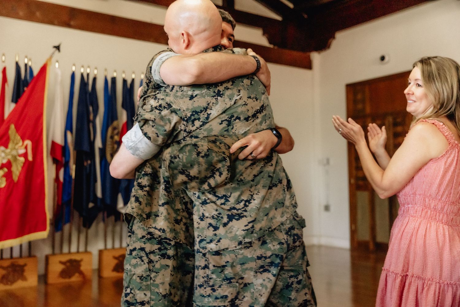 Two Marines embrace warmly at a military ceremony while a smiling woman in pink applauds nearby.