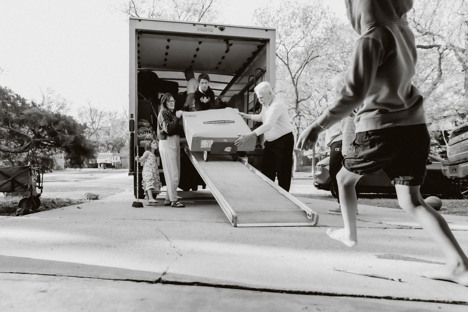People unloading furniture from a moving truck using a ramp in a residential neighborhood.