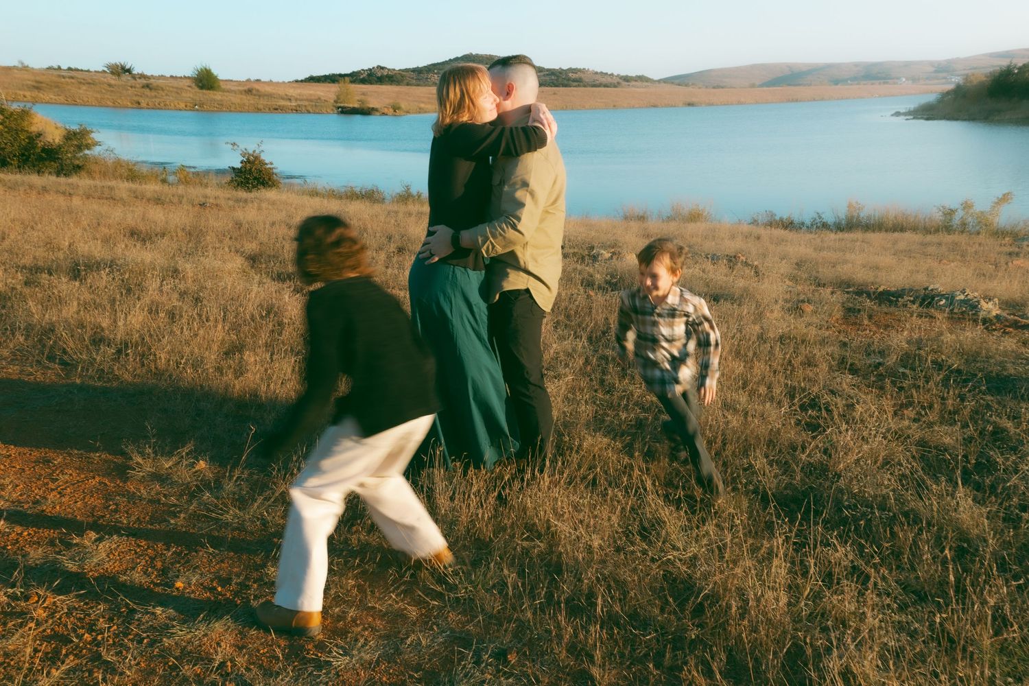 Family embracing in golden field near a lake, with two children playing around them on a sunny day.