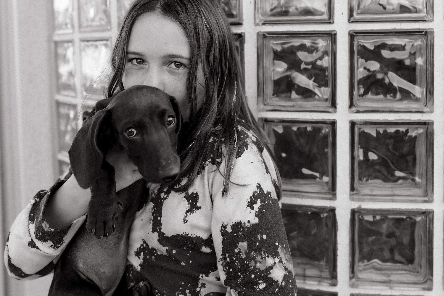 Black and white photo of a young girl holding a dachshund puppy close to her face near a glass block window.