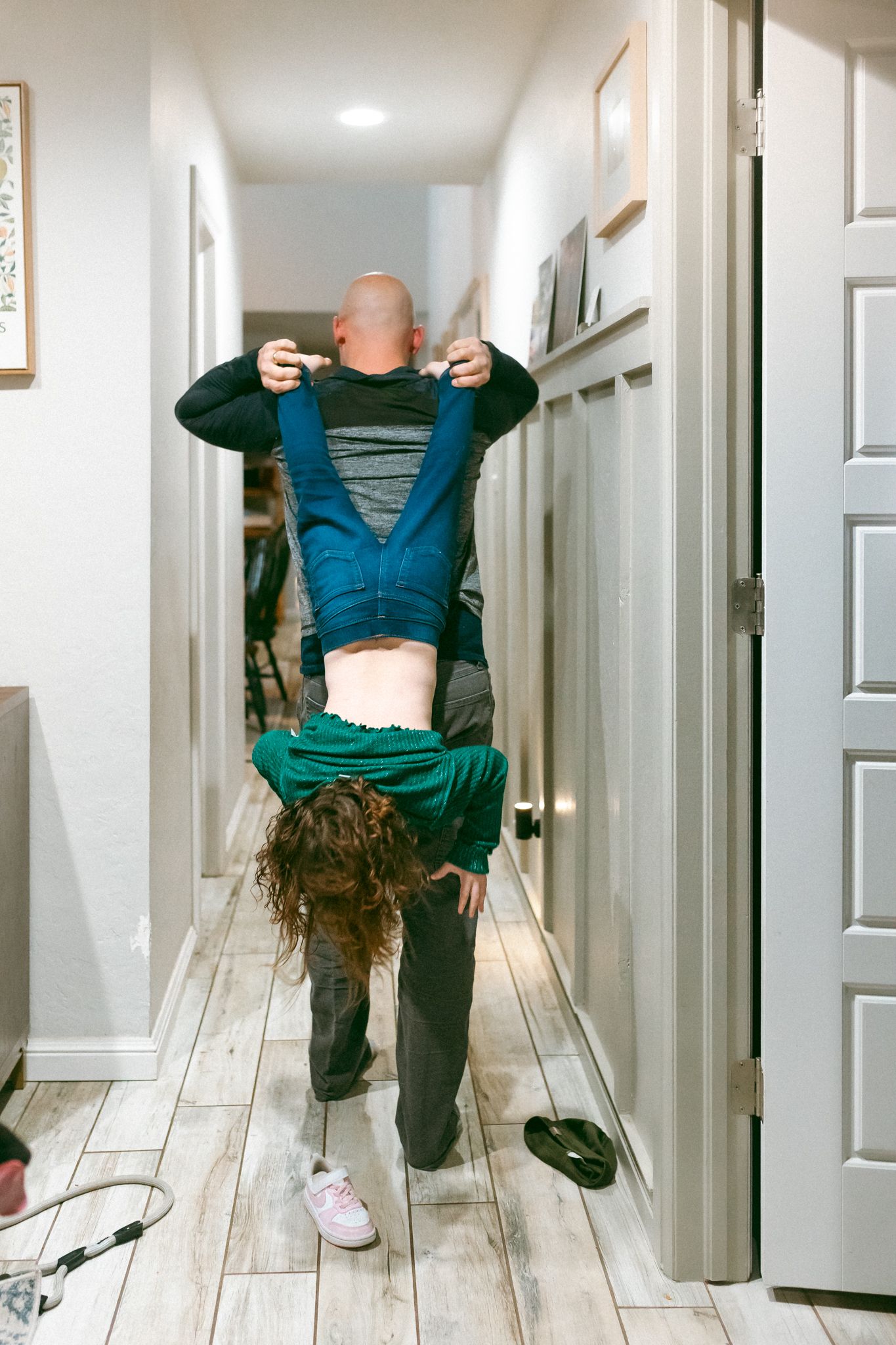 A person wearing a green sweater and blue overalls stands upside down in a narrow hallway with white walls.