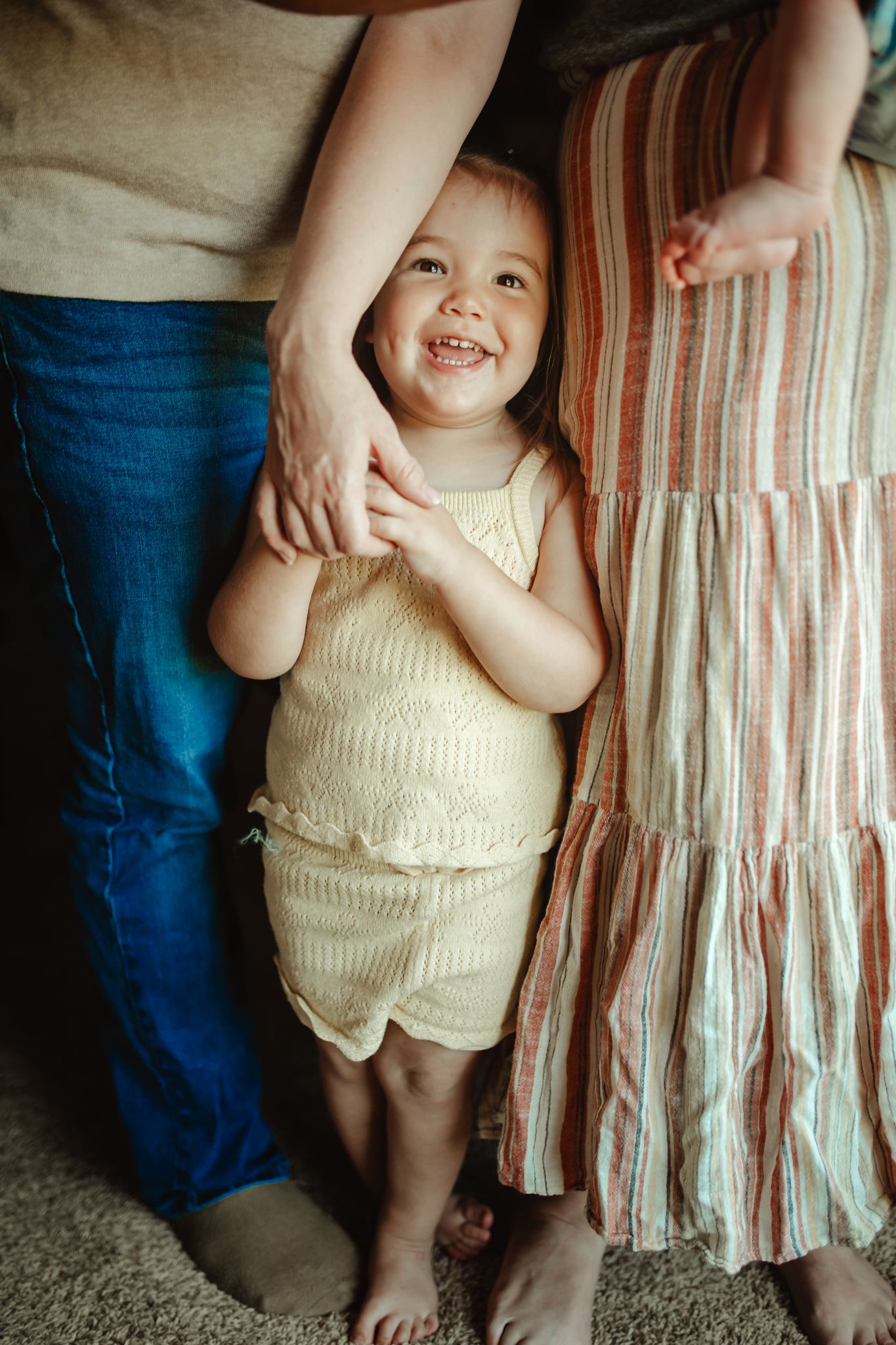 A small child wearing a cream-colored knit romper stands against a striped curtain background.