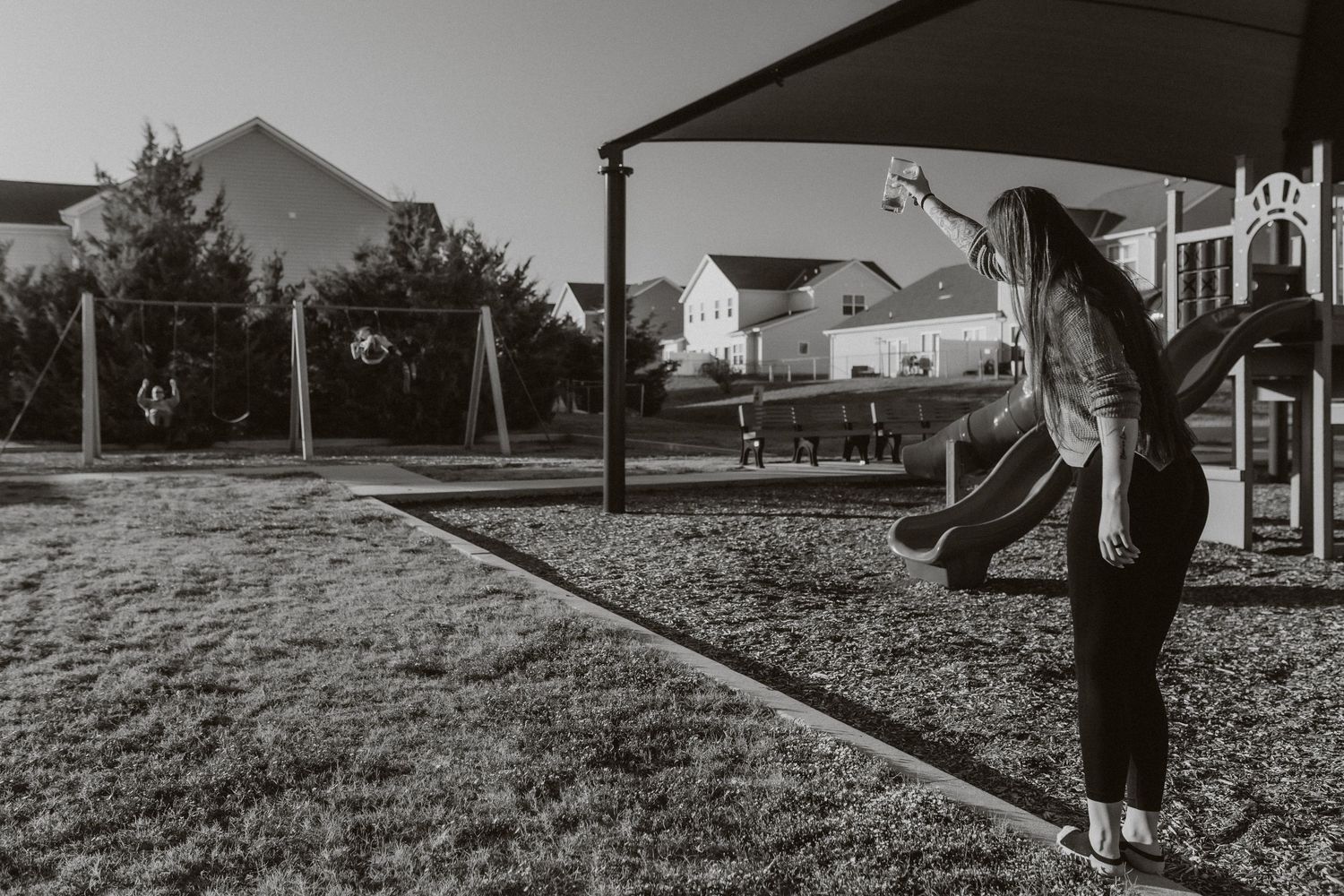 Person on playground swings in black and white photograph with houses in background.