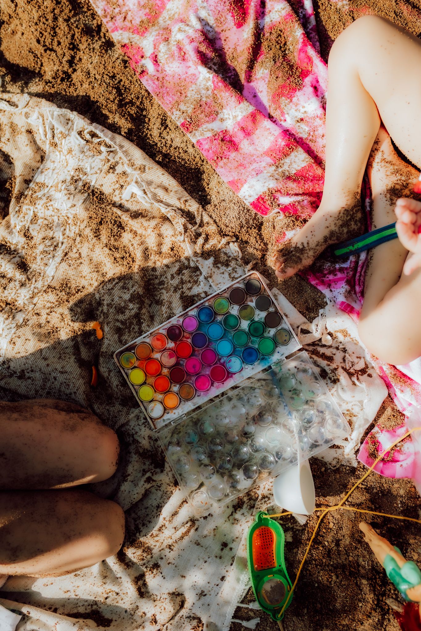 Colorful watercolor paints and art supplies scattered on sandy ground during outdoor activity.