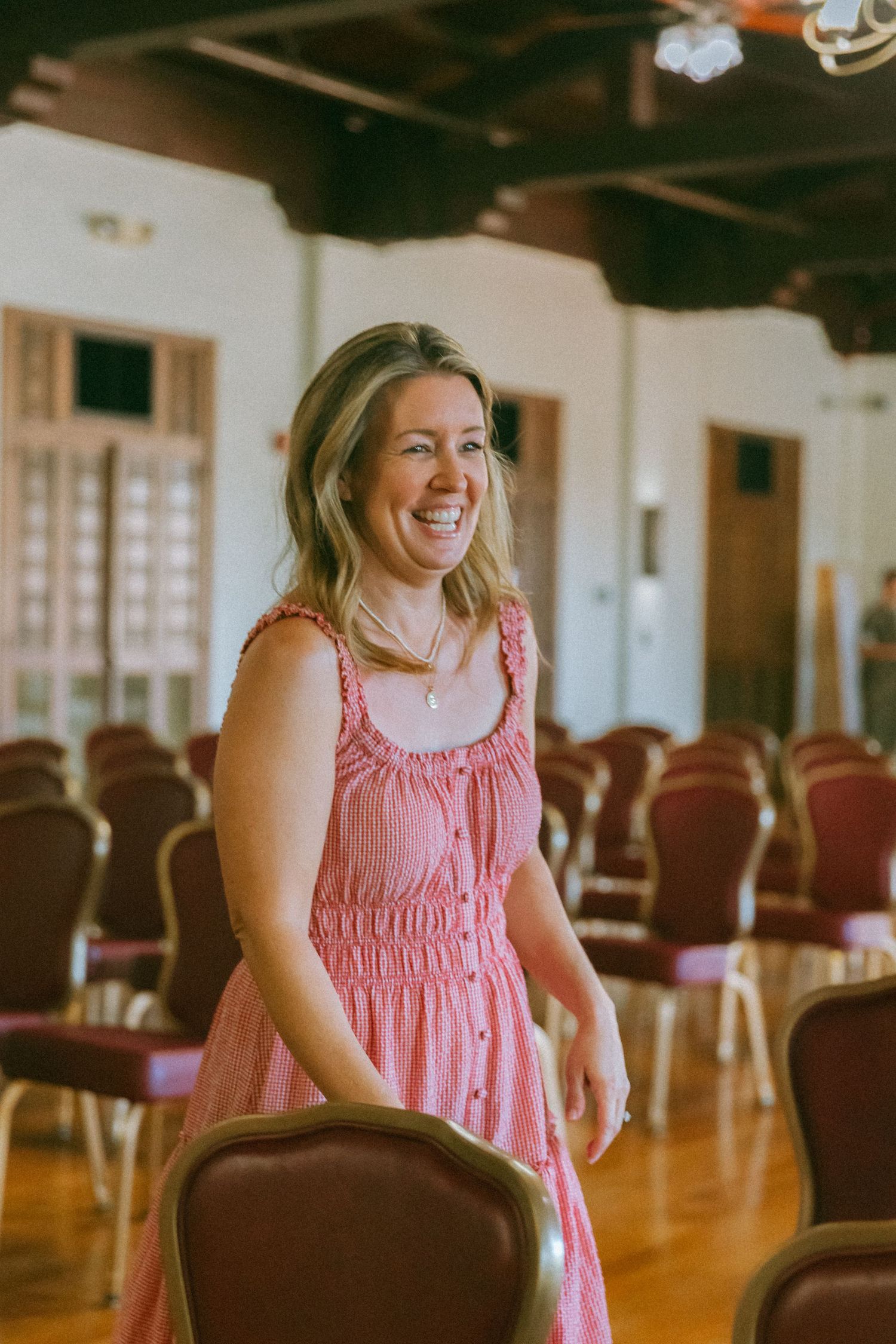 Smiling woman in a pink dress standing among rows of red chairs in an elegant hall.