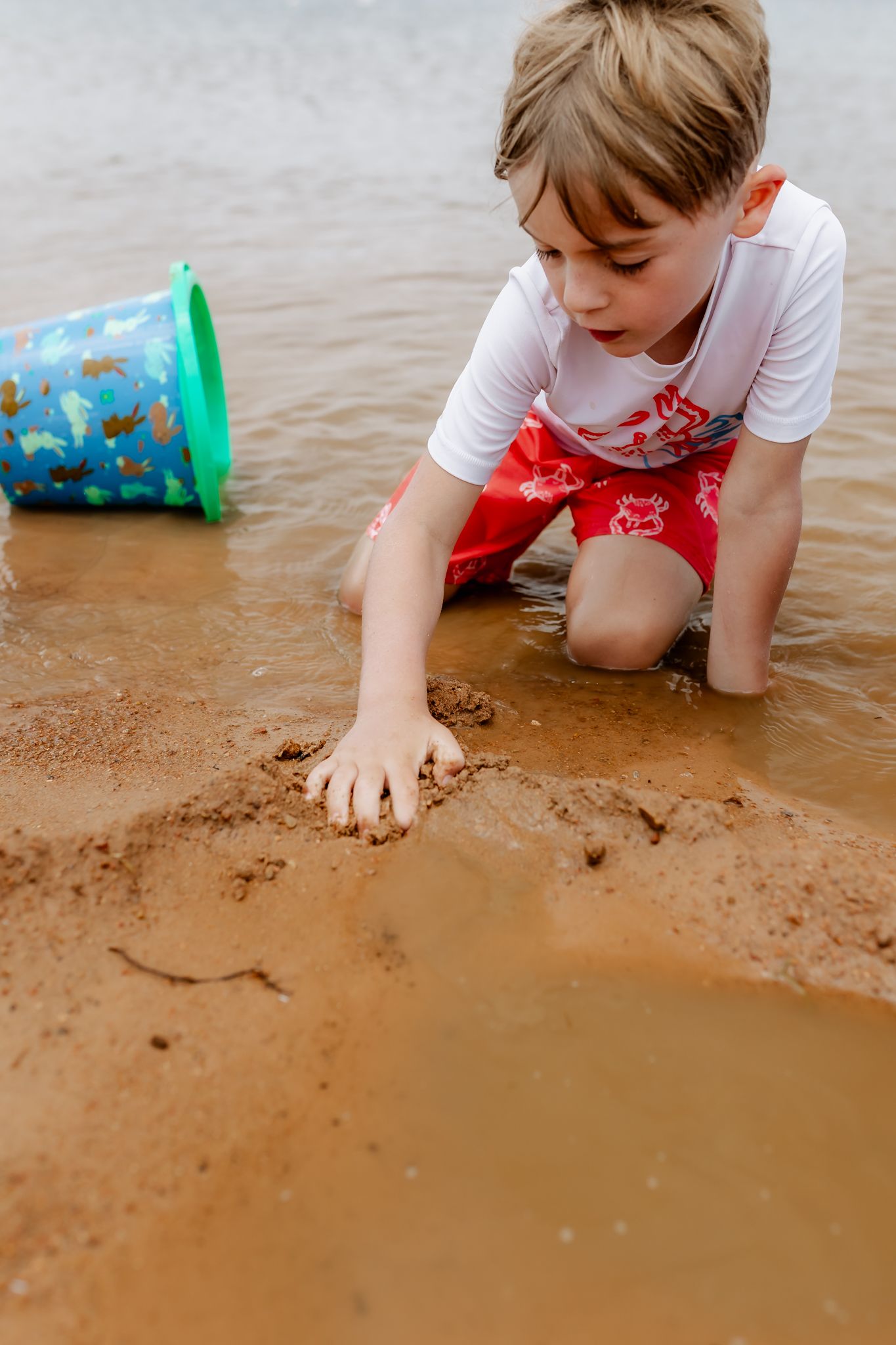 Young child in white shirt playing with wet sand near a colorful bucket at the water's edge.