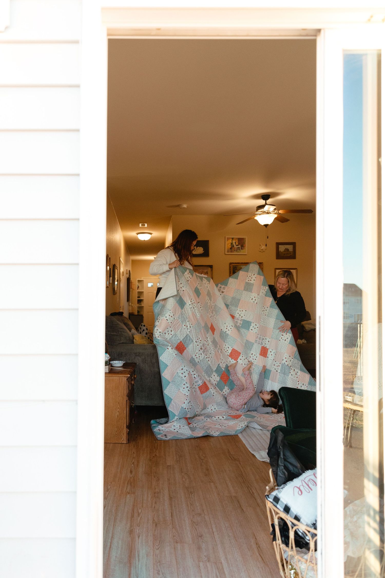 A handmade quilt with blue and red patterns is being held up in a sunlit living room with hardwood floors.