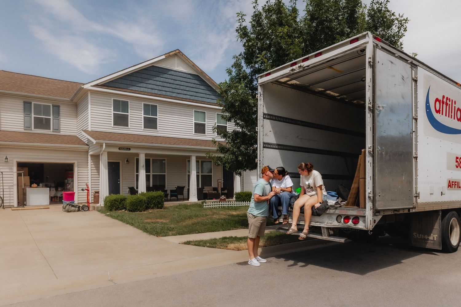 People sitting on the back of an Affinity moving truck parked outside a suburban home during a move.