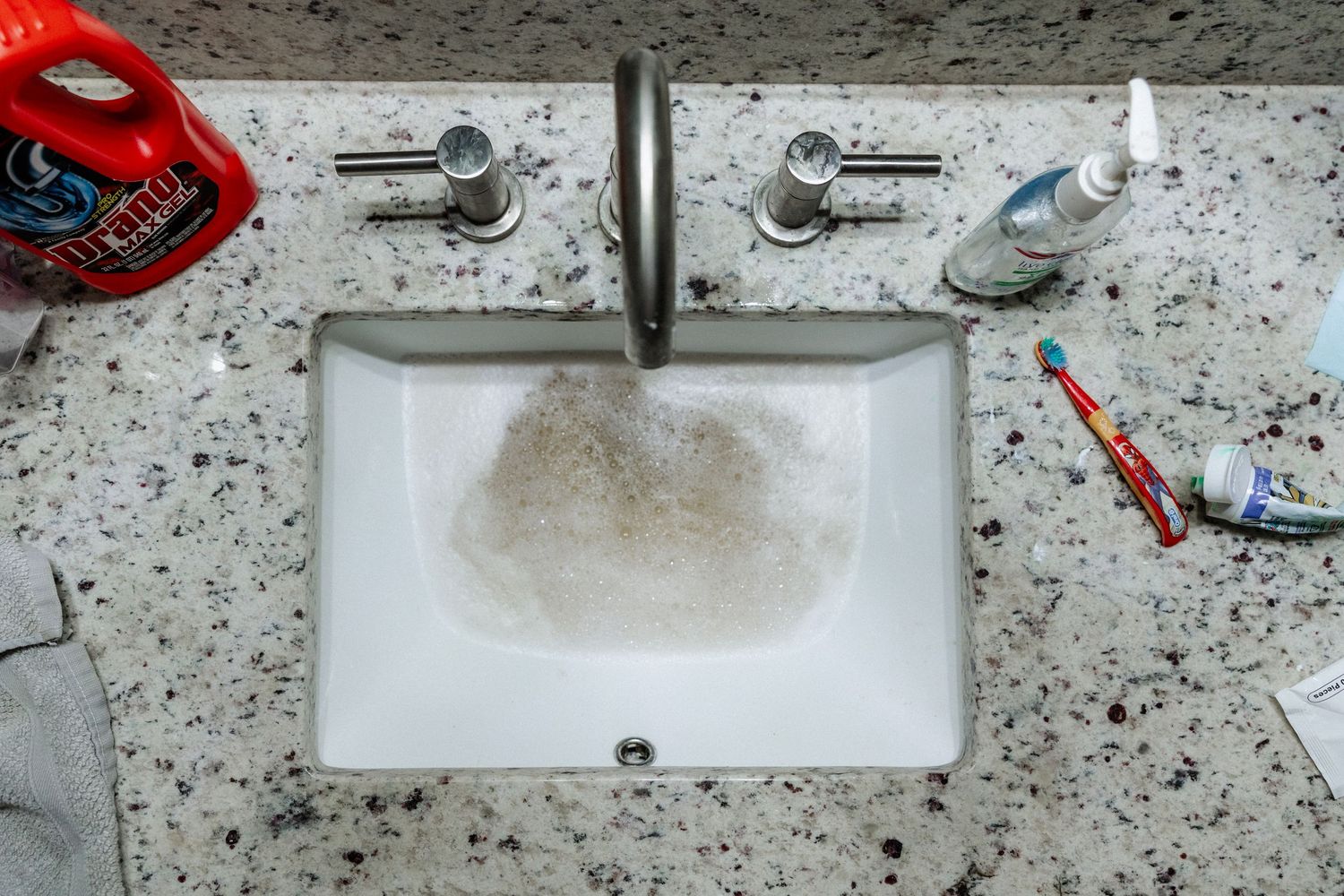 Overhead view of a white bathroom sink with running water on a granite countertop with toothbrush and toiletries and mom's duties to unclog