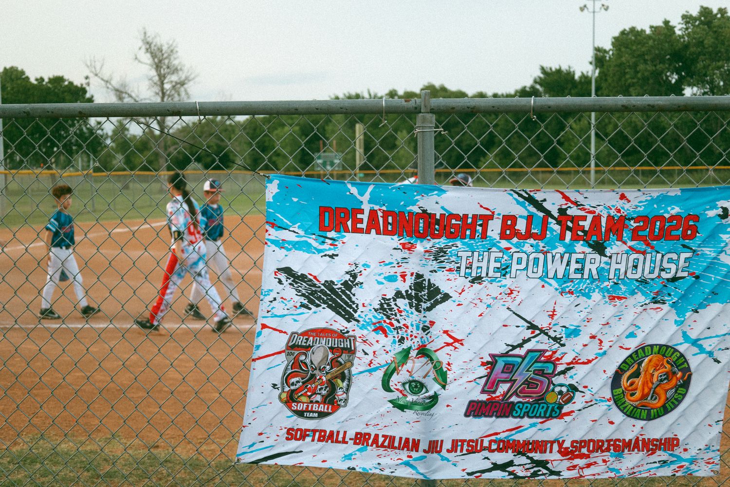 Youth baseball players on field near Dreamnight BJJ Team 2026 The Power House banner at outdoor diamond.