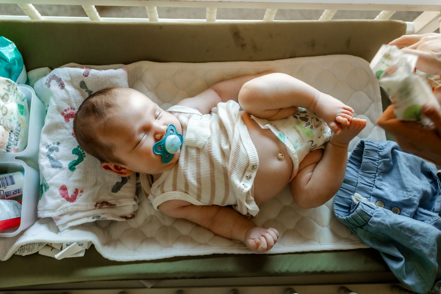 A baby in a striped onesie with pacifier resting on a green couch surrounded by clothing.