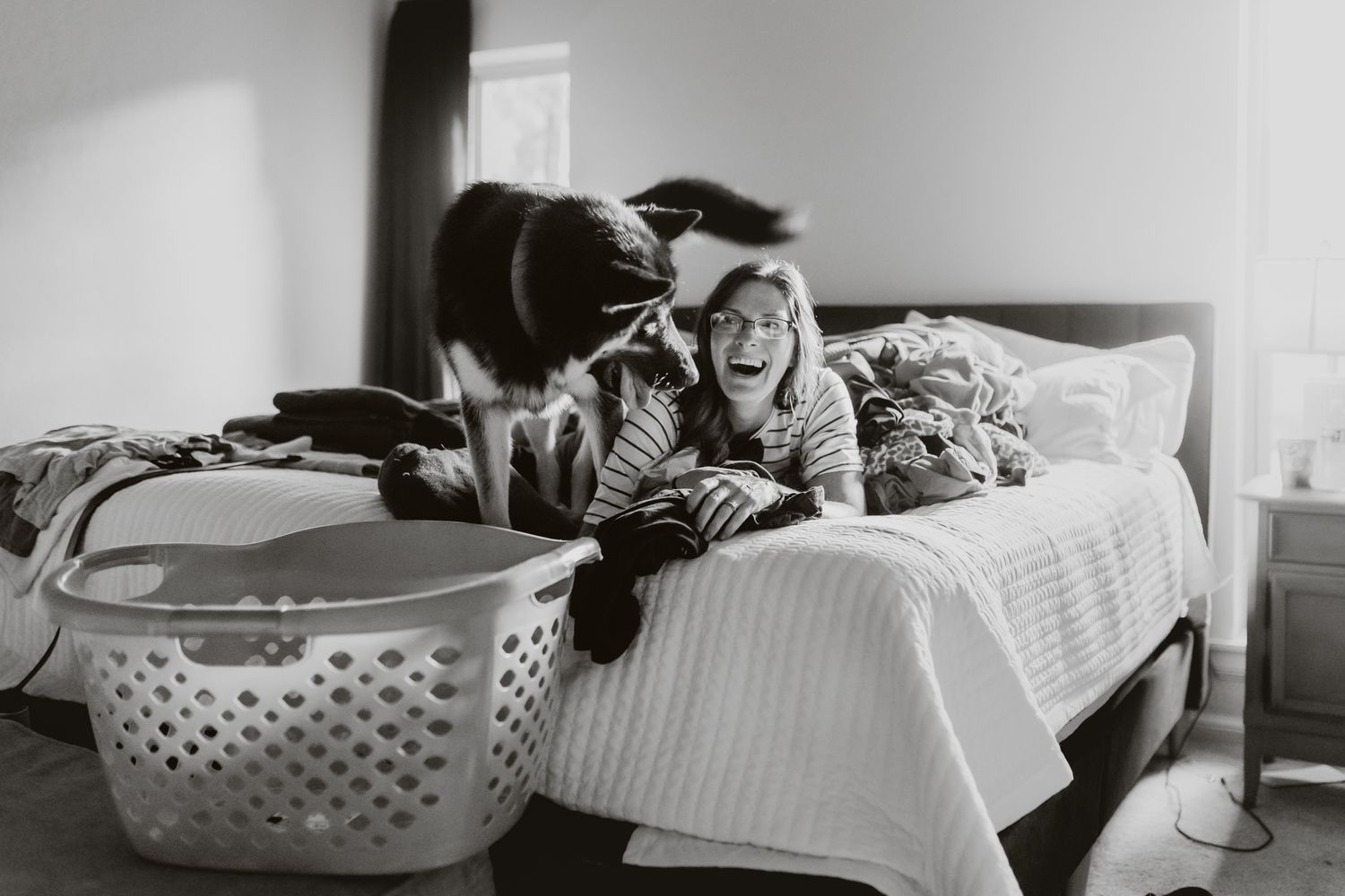 Black and white photo of a cozy bedroom scene with a cat relaxing on a quilted bed near a laundry basket.