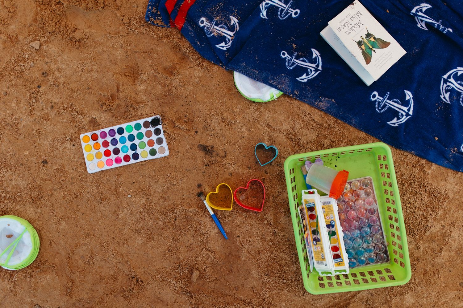 Beach toys and watercolor set scattered on sandy ground with anchor pattern fabric.