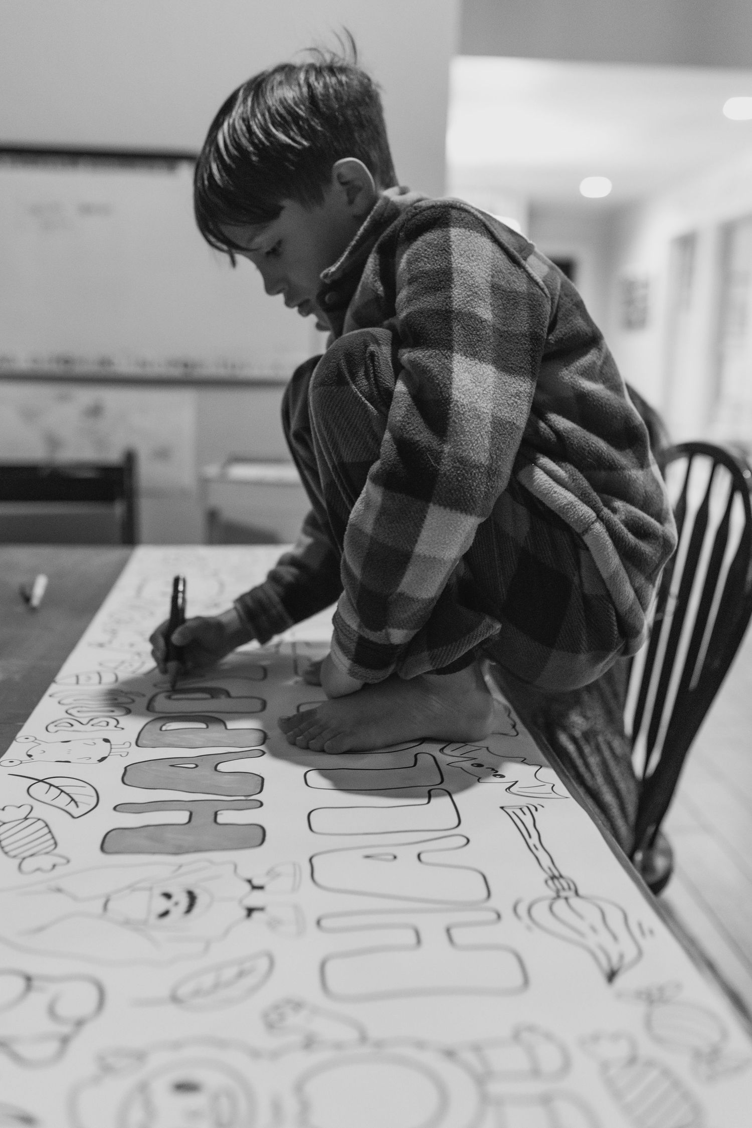 Boy in plaid shirt coloring a Halloween-themed paper banner at a table in black and white photo.