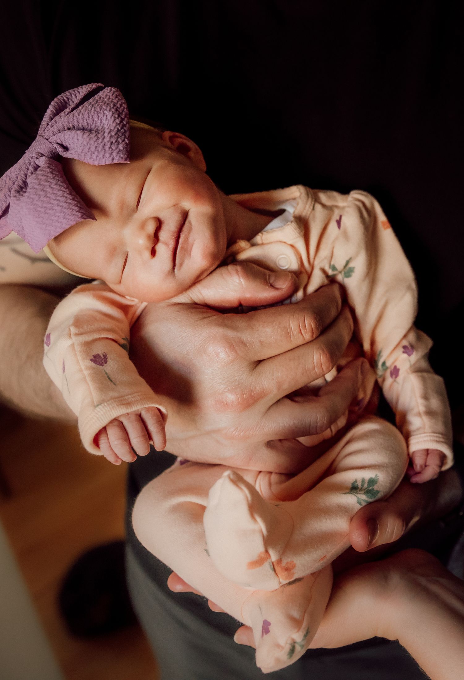 Newborn baby in floral onesie and purple bow headband sleeping peacefully while being cradled in adult hands.
