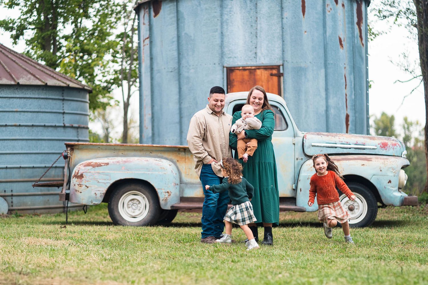 Family posing by a vintage blue pickup truck on a farm, parents holding baby while children play outdoors during their session in Centerview
