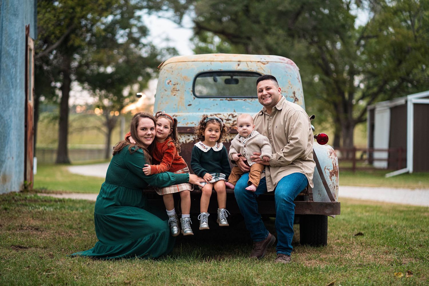 A family sits on the back of a blue truck and smiles at the camera during their family photography session located in Lee's Summit