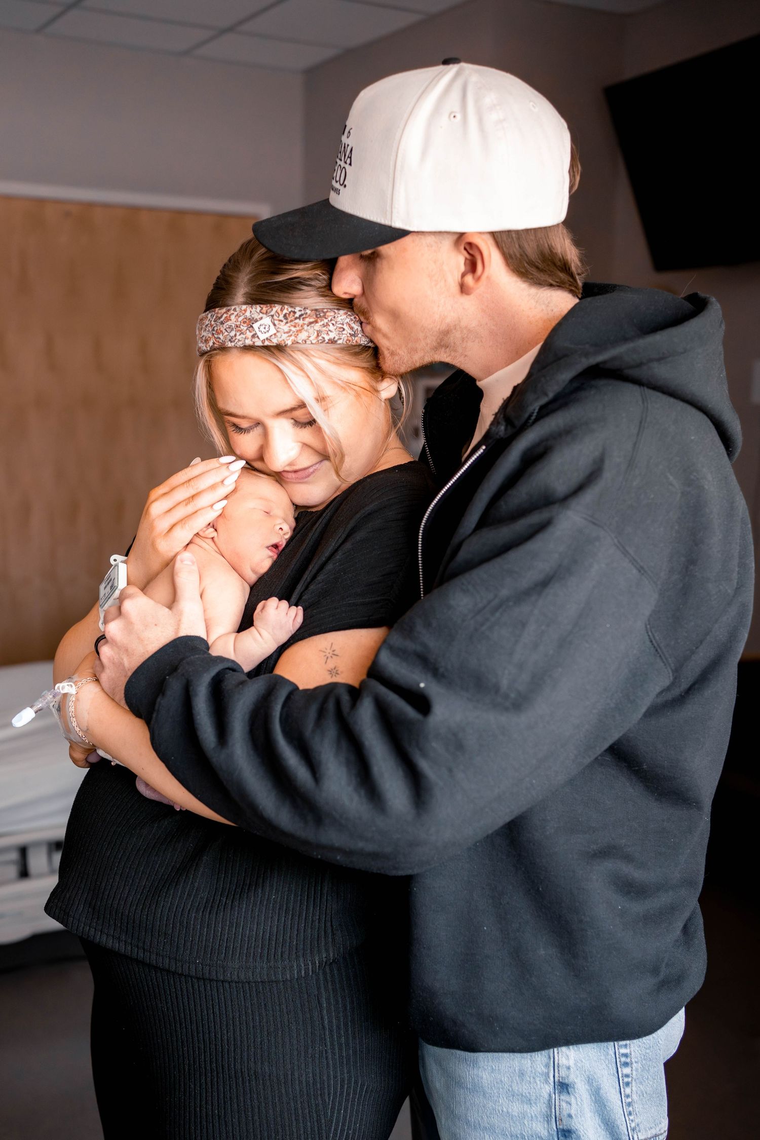 New parents share a tender moment, with father kissing mothers forehead as she cradles their newborn baby in a hospital room.