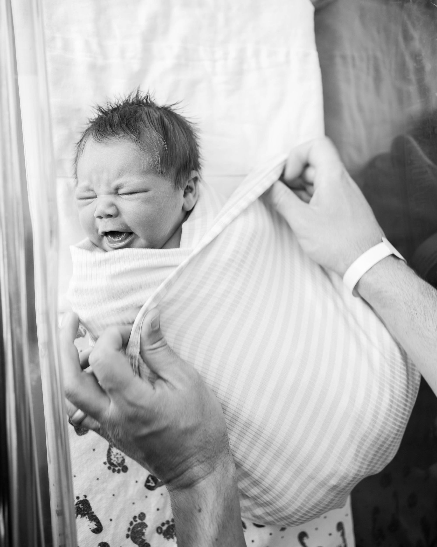 Black and white photo of a crying newborn baby swaddled in a striped blanket, held by adult hands in a hospital.