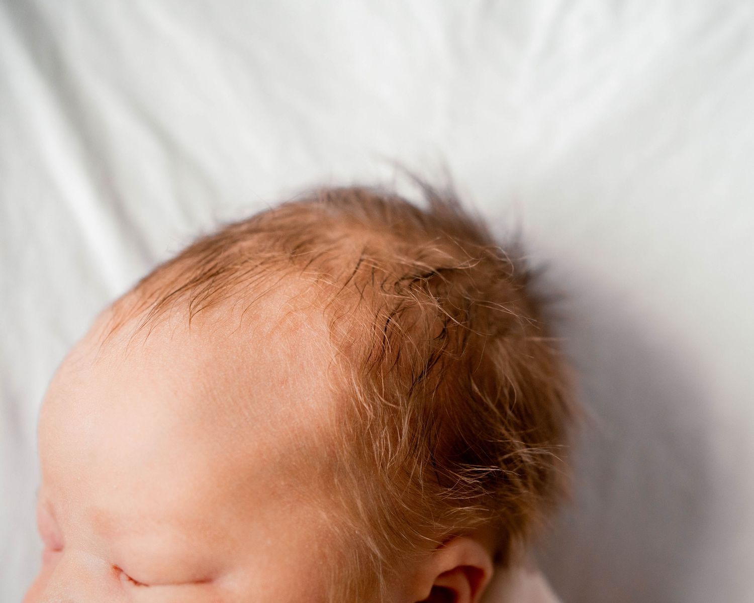 Close-up of a sleeping newborn baby with fine reddish hair on a soft white background.