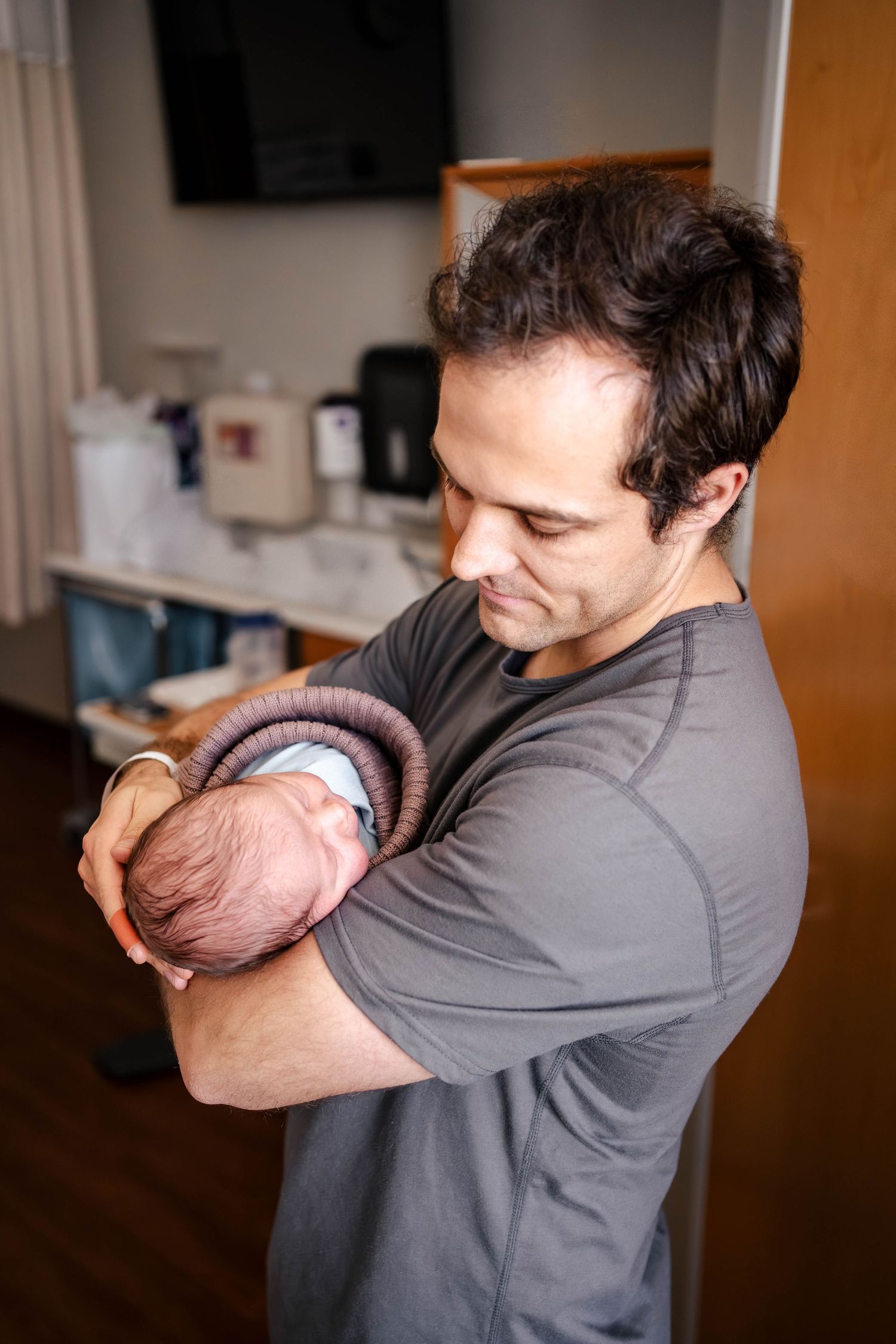 A father tenderly holds his newborn baby in a hospital room, gazing down with a warm smile.
