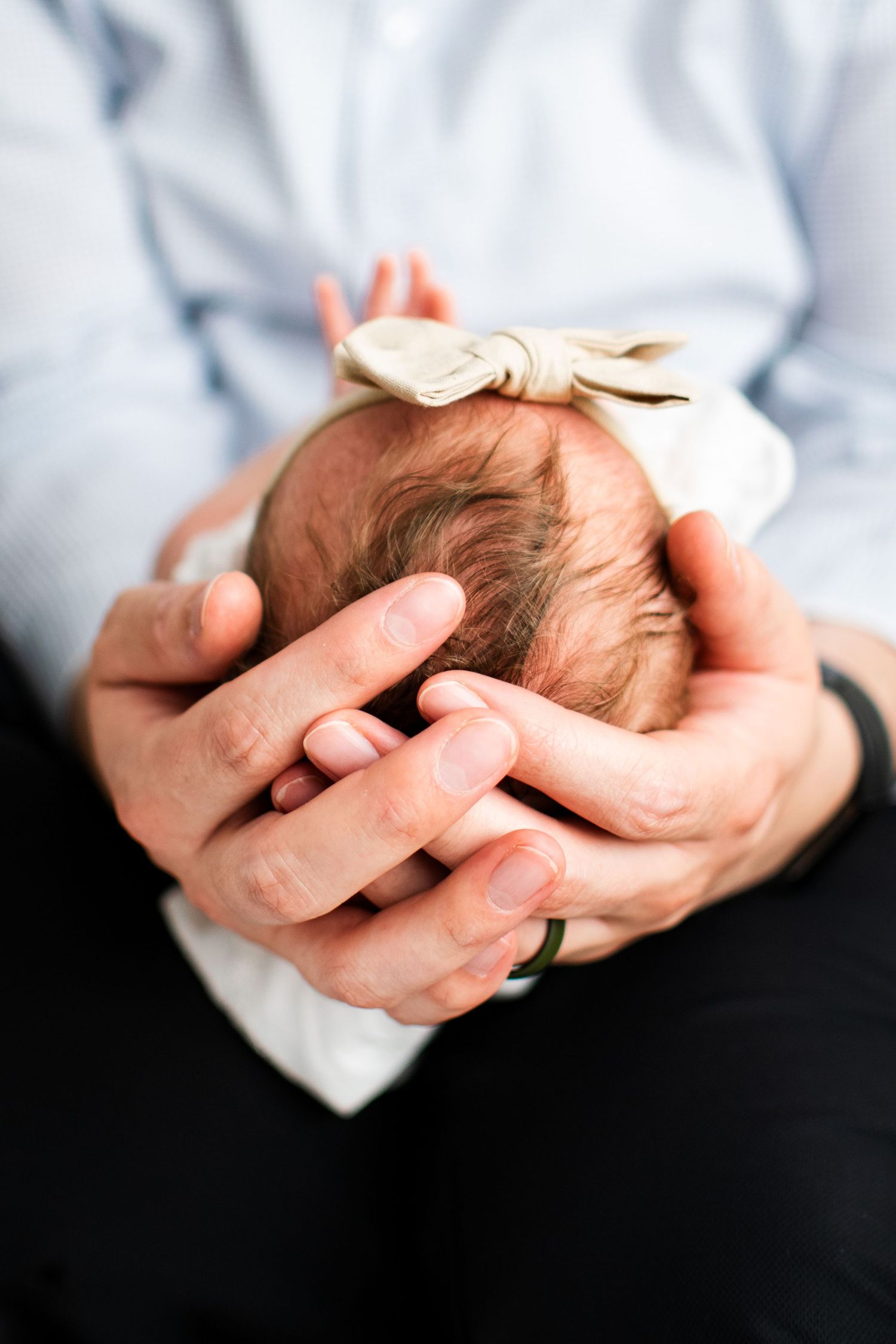 Gentle hands cradle a newborn baby wearing a soft bow headband in an intimate close-up portrait.