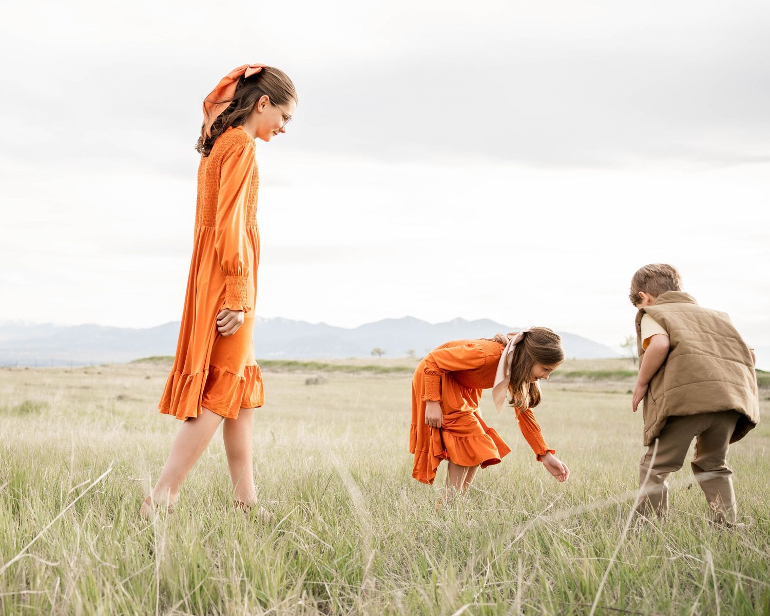 Three children in orange and earth-toned outfits explore a tall grass meadow with distant mountains under a soft, overcast sky.