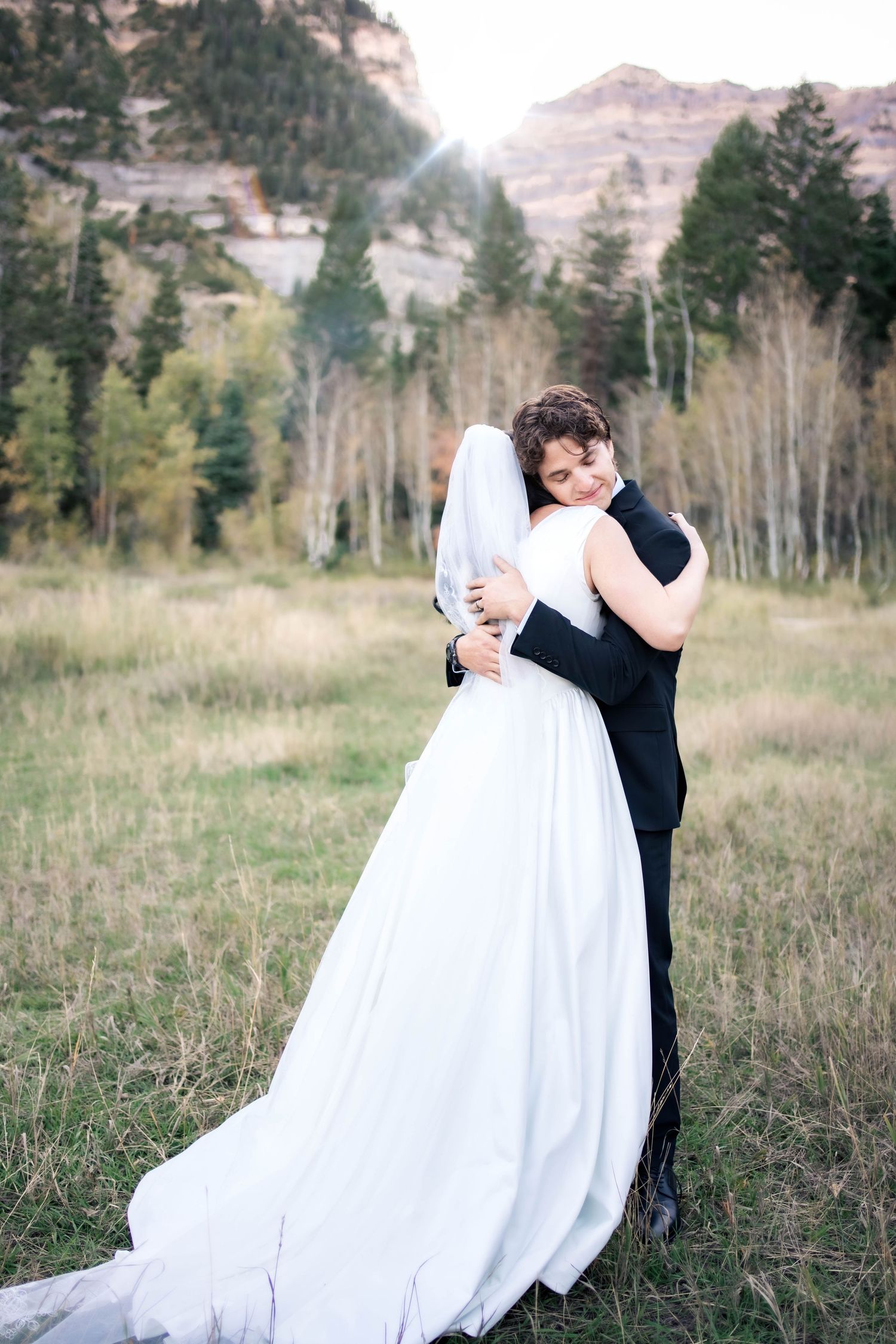 Bride in a flowing white gown and veil embraces a groom in a dark suit in a grassy mountain meadow with golden aspen trees at sunset.
