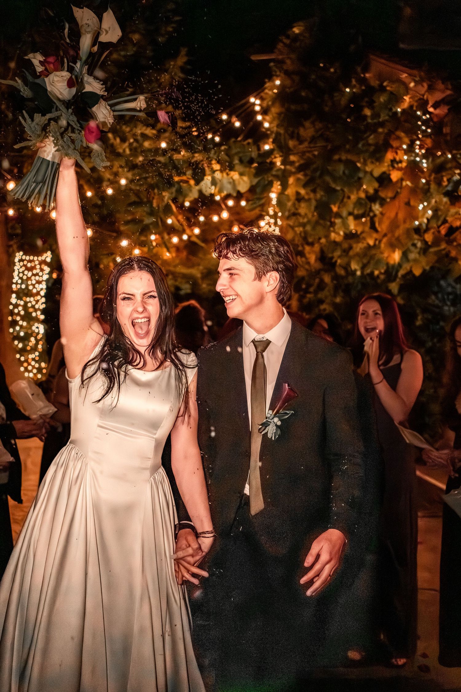 Joyful bride in a satin gown raises her bouquet while holding hands with the groom at a night wedding under glowing string lights.