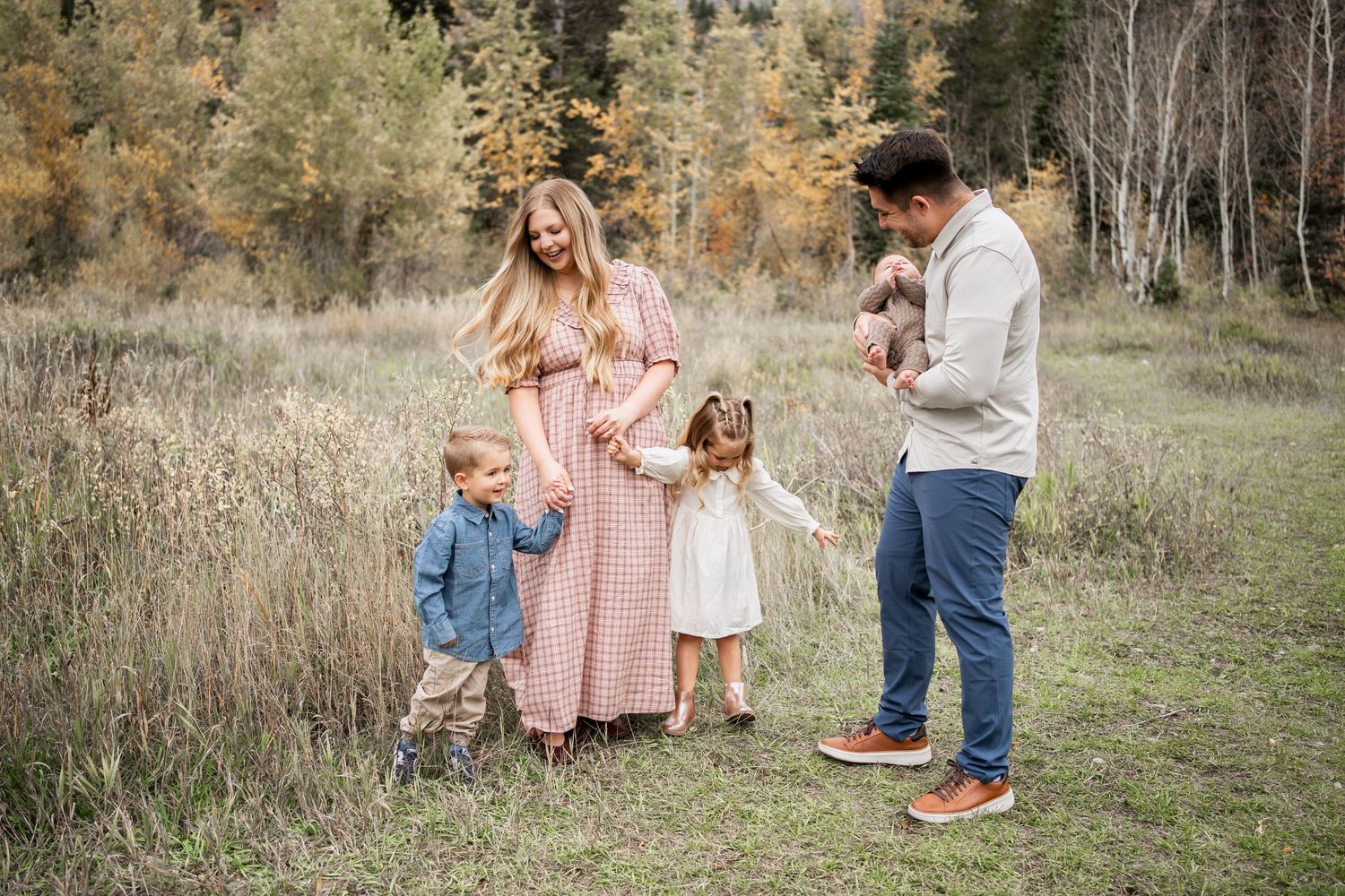 Family of four in an autumn field, with a mother in a pink dress, a father holding a baby, and two young children holding hands.