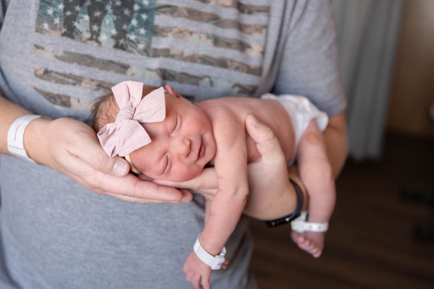 Newborn baby wearing a pink bow headband and hospital ID bracelets rests peacefully in the cradled hands of a person in a gray t-shirt.