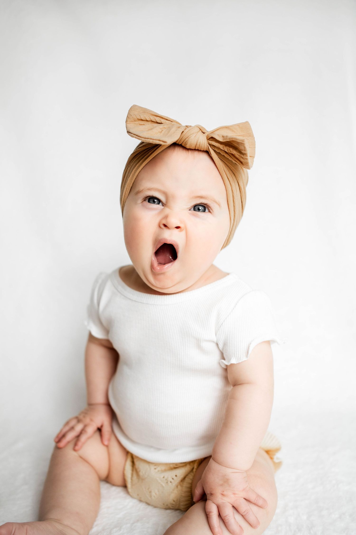 Adorable baby girl in white onesie and tan bow headband sitting with mouth wide open in surprise.