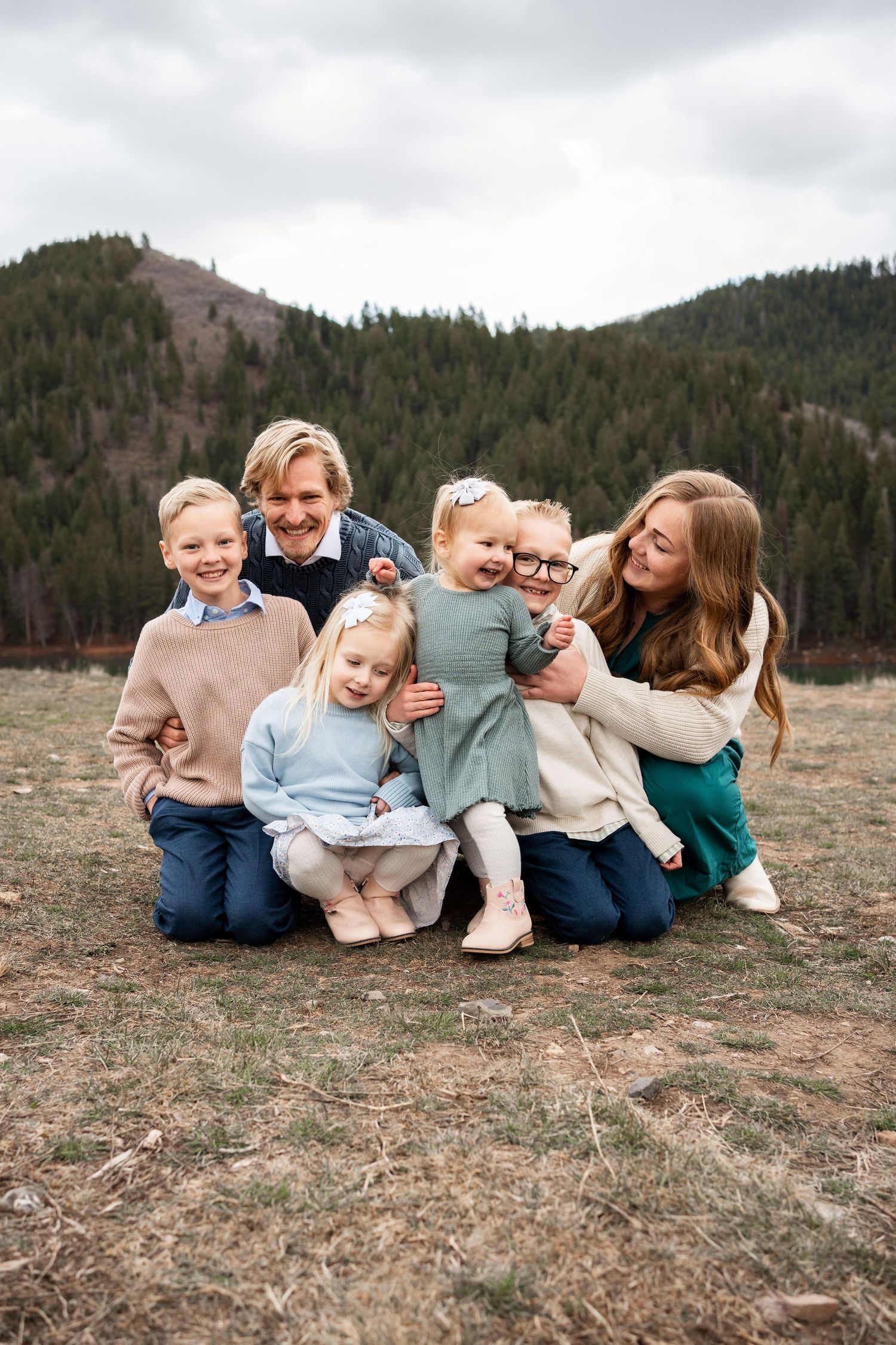 A family group lounges together on a grassy hillside with mountain views in casual coordinated outfits.